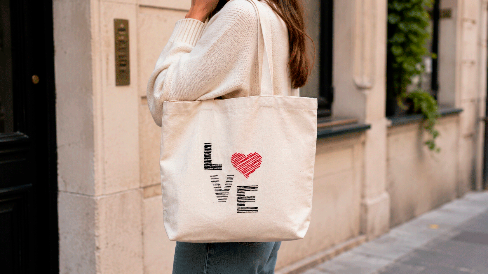 Woman carrying a cream cotton tote bag embroidered with the LOVE design in black, gray, and red thread over her shoulder while walking through the city.