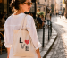 Woman in a white shirt carrying a handmade LOVE embroidered tote bag through a sunlit cobblestone street, showing how a simple embroidery project becomes an everyday accessory.