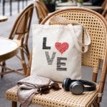 Natural cotton LOVE tote bag with black embroidery and a red heart on a woven café chair