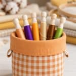 Six wooden embroidery bobbins wound with orange, purple, gold, beige, brown and green floss in an orange gingham fabric basket