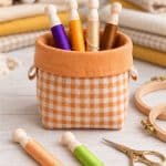Four wooden embroidery bobbins in a gingham basket, with two more bobbins, gold scissors and wooden hoops on a white table