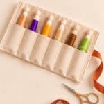Six wooden thread bobbins in autumn colors tucked into a coton canvas roll-up pouch, with gold embroidery scissors and an orange ribbon